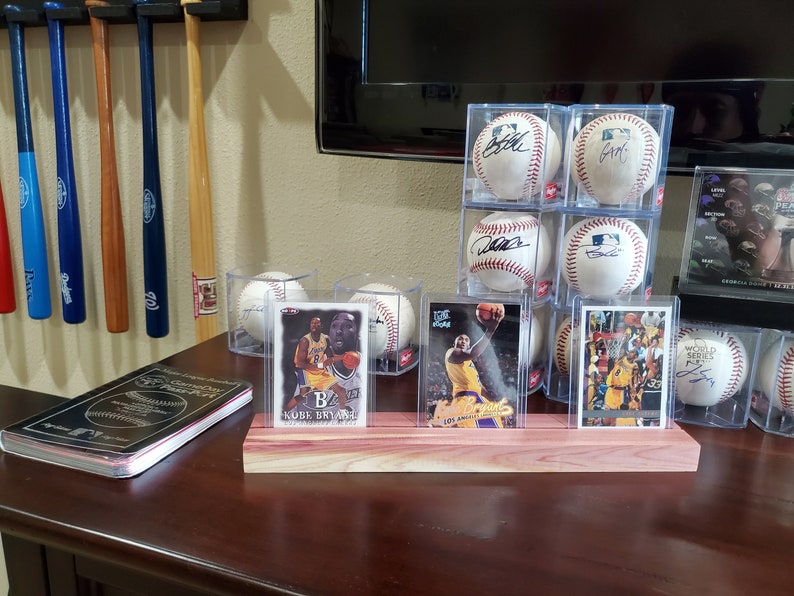May include: Three clear plastic display cases with signed baseballs and three clear plastic display cases with basketball trading cards featuring Kobe Bryant. The display cases are on a wooden stand.