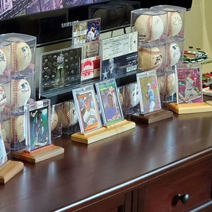 May include: A collection of baseball cards and signed baseballs displayed on a wooden shelf. The cards are in protective cases and the baseballs are in clear plastic domes. The shelf is brown wood with a drawer below.