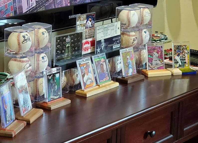 May include: A collection of baseball cards and signed baseballs displayed on a wooden shelf. The cards are in protective cases and the baseballs are in clear plastic domes. The shelf is brown wood with a drawer below.