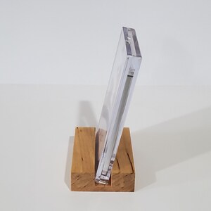 May include: A clear plastic display stand with a wooden base. The stand is designed to hold a CD or DVD case.