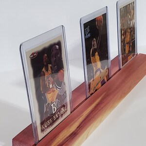May include: Three basketball trading cards in protective plastic cases are displayed on a wooden stand. The cards feature Kobe Bryant, a basketball player for the Los Angeles Lakers.