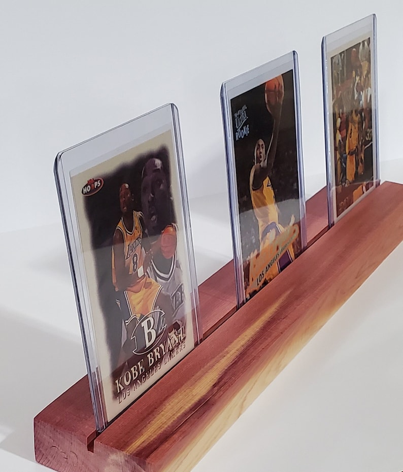 May include: Three basketball trading cards in protective plastic cases are displayed on a wooden stand. The cards feature Kobe Bryant, a basketball player for the Los Angeles Lakers.