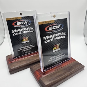 以下が含まれることがあります： 「BCW Magnetic Card Holder」の文字が入った、木製の台座に置かれた2つの透明なマグネット式カードホルダー。 台座は長方形で、濃い茶色の仕上げと赤いアクセント。 35ptカード用です。