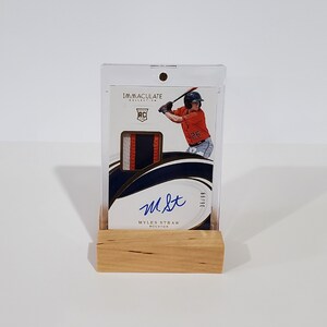 May include: A baseball card featuring a baseball player in a white and orange uniform, holding a bat. The card is in a clear plastic case with a wooden base. The card is signed "M.St." and has the text "Myles Straw Houston" and "Immaculate Collection" printed on it.
