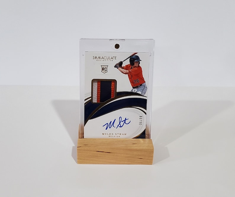 May include: A baseball card featuring a baseball player in a white and orange uniform, holding a bat. The card is in a clear plastic case with a wooden base. The card is signed "M.St." and has the text "Myles Straw Houston" and "Immaculate Collection" printed on it.