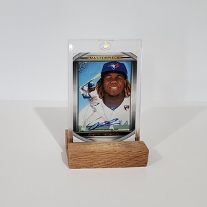 May include: A baseball card in a clear plastic case with a wooden stand. The card features a baseball player in a blue uniform with a white jersey. The card has a gold border and the word "MASTERPIECE" printed on it.
