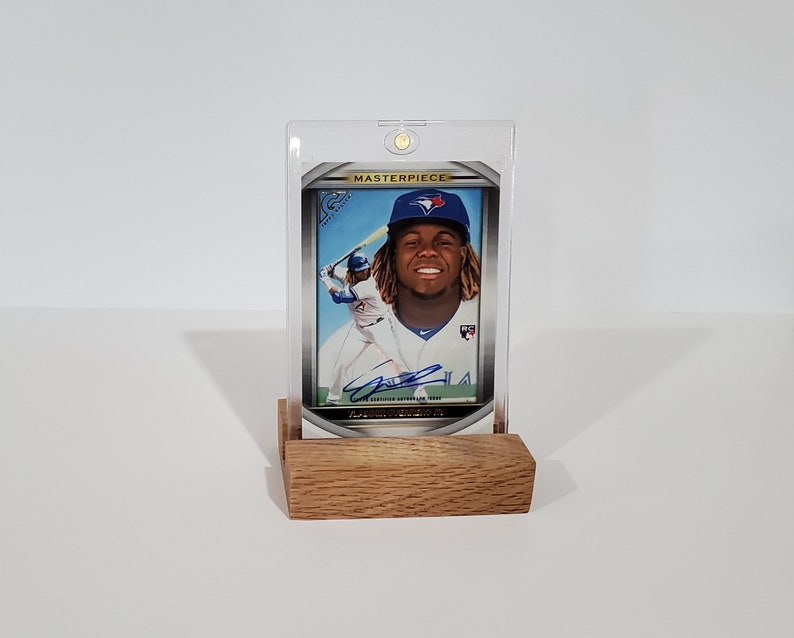 May include: A baseball card in a clear plastic case with a wooden stand. The card features a baseball player in a blue uniform with a white jersey. The card has a gold border and the word "MASTERPIECE" printed on it.