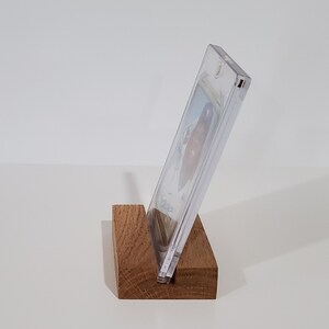 May include: A clear plastic display case with a card inside, resting on a wooden stand. The stand is a dark brown color and has a simple design.