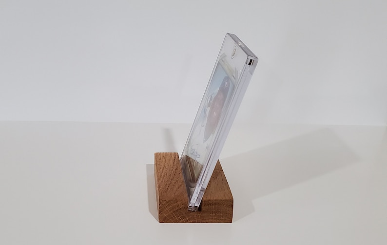 May include: A clear plastic display case with a card inside, resting on a wooden stand. The stand is a dark brown color and has a simple design.