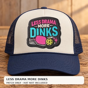May include: A navy blue and cream trucker hat featuring an embroidered patch on the front. The patch displays the text LESS DRAMA MORE DINKS in pink and blue, accompanied by a pink pickleball paddle and a yellow pickleball with small stars.
