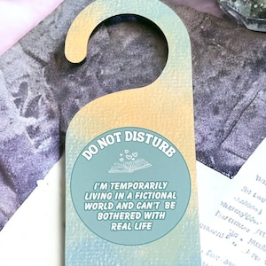 May include: A teal and yellow door hanger with a white circle that says "DO NOT DISTURB" and "I'M TEMPORARILY LIVING IN A FICTIONAL WORLD AND CAN'T BE BOTHERED WITH REAL LIFE". The circle has a white open book with three hearts above it.