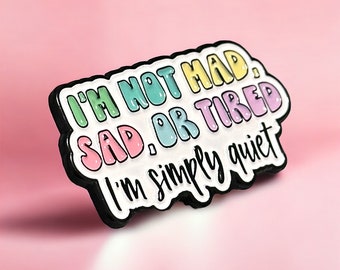 May include: A white enamel pin with black outlining. The pin says "I'm not mad, sad, or tired. I'm simply quiet." The words are in different colors: green, pink, blue, and purple.