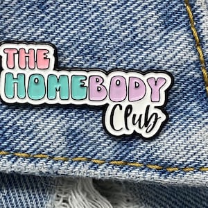 May include: A colorful enamel pin with the text "THE HOMEBODY CLUB" in pink, teal, and purple, outlined in black. The pin is set against a blue denim background.
