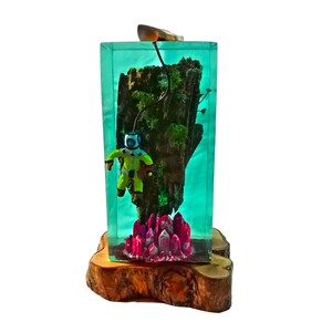 May include: A small green figurine of a scuba diver is encased in a clear resin block with a piece of driftwood and pink crystals. The block is sitting on a wooden base.