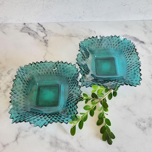 May include: Two teal blue glass square dishes with a diamond pattern. The dishes are on a white marble surface.