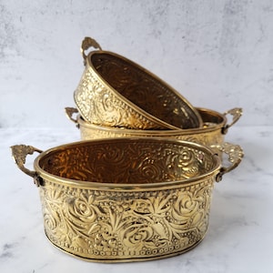 May include: A set of three gold-colored, oval-shaped metal bowls. Each bowl features an embossed floral design and decorative handles. The bowls are stacked, with the largest bowl in the foreground.