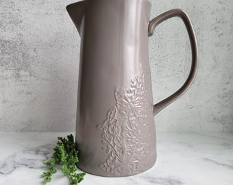 May include: A gray ceramic pitcher with an embossed floral design. The pitcher has a wide mouth and a handle.