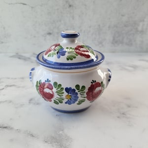 May include: A white ceramic sugar bowl with a lid, decorated with hand-painted blue, red, and green floral designs.