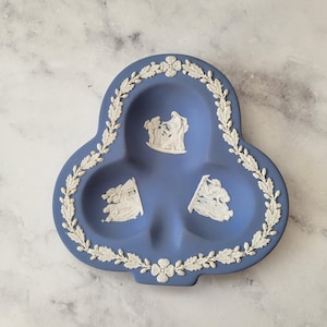 May include: A blue ceramic trinket dish with three compartments and a white floral and leaf border. The dish features white relief figures in each compartment, showcasing a classical theme.