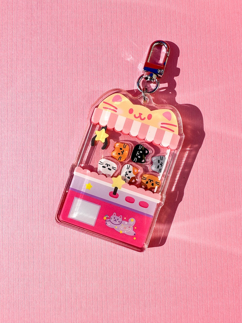 Peut inclure: Un porte-cl&eacute;s en plastique transparent en forme de machine &agrave; griffes sur le th&egrave;me du chat avec un design rose et violet. La machine &agrave; griffes a une &eacute;toile, un visage de chat et une patte de chat sur le dessus. Il y a six petits visages de chat &agrave; l'int&eacute;rieur de la machine &agrave; griffes. Le porte-cl&eacute;s a un anneau en m&eacute;tal argent&eacute;.