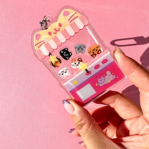Peut inclure: Un porte-cl&eacute;s en forme de chat rose et jaune avec une fen&ecirc;tre en plastique transparent. La fen&ecirc;tre montre une vari&eacute;t&eacute; de visages de chats color&eacute;s &agrave; l'int&eacute;rieur d'une machine &agrave; griffes de jouet. Le porte-cl&eacute;s a un anneau en m&eacute;tal argent&eacute;.