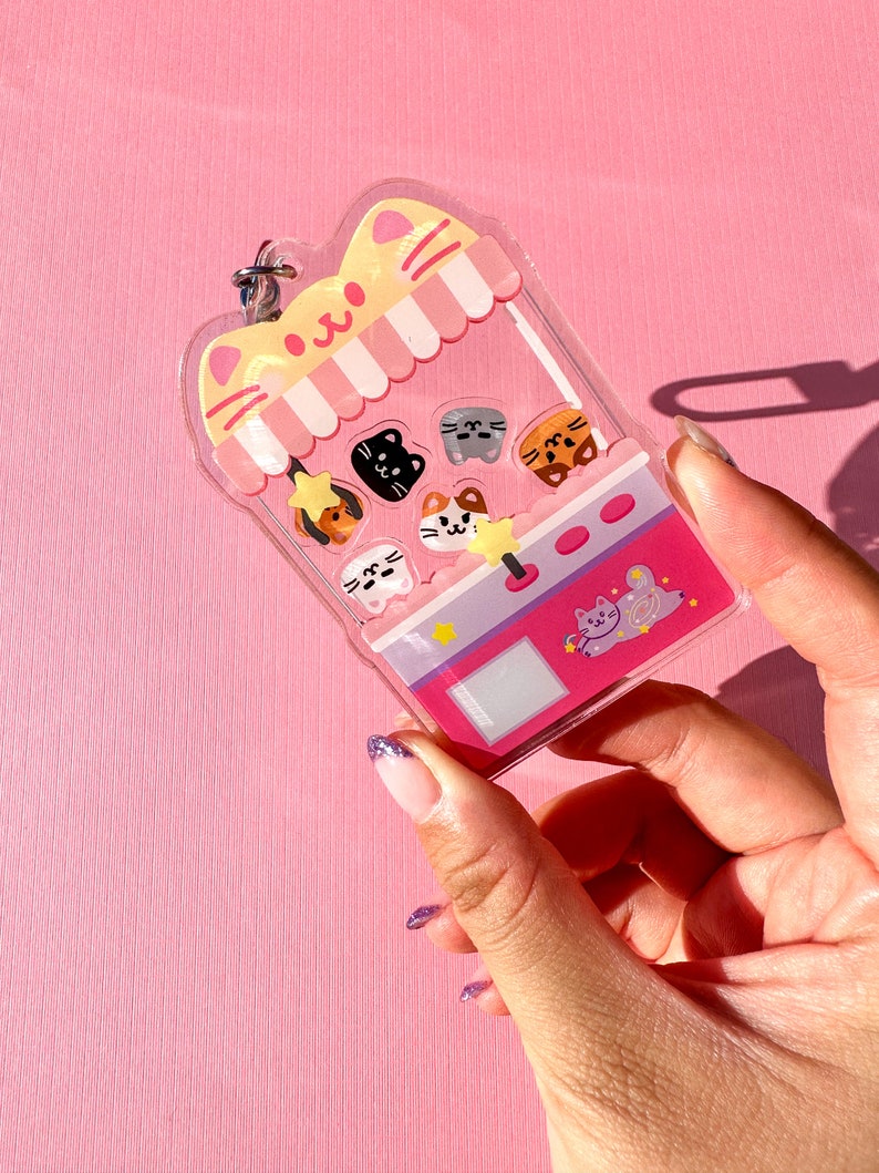 Peut inclure: Un porte-cl&eacute;s en forme de chat rose et jaune avec une fen&ecirc;tre en plastique transparent. La fen&ecirc;tre montre une vari&eacute;t&eacute; de visages de chats color&eacute;s &agrave; l'int&eacute;rieur d'une machine &agrave; griffes de jouet. Le porte-cl&eacute;s a un anneau en m&eacute;tal argent&eacute;.