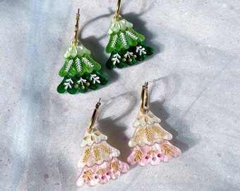 May include: Three pairs of Christmas tree-shaped earrings. Two pairs are green and white, and one pair is pink and white. The earrings have gold-colored hoops and are decorated with gold and red accents. The earrings are on a white, sparkly surface.