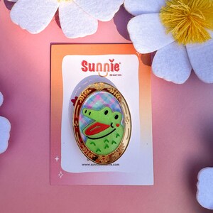 May include: A green alligator with a red mouth and a pink heart on its cheek is featured on a gold oval-shaped pin. The pin is surrounded by a gold frame with a floral design. The pin is on a white card with the text "Sunnie Creative" and a website address.