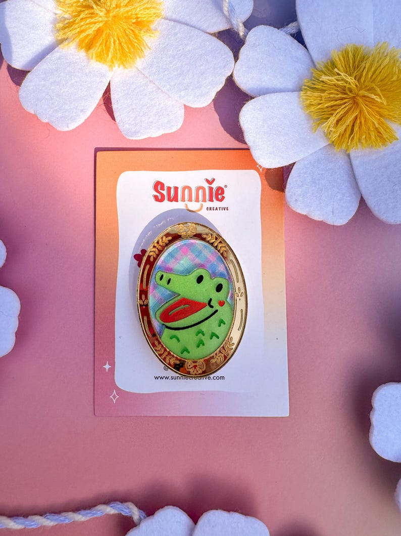 May include: A green alligator with a red mouth and a pink heart on its cheek is featured on a gold oval-shaped pin. The pin is surrounded by a gold frame with a floral design. The pin is on a white card with the text "Sunnie Creative" and a website address.