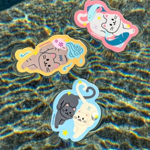 May include: Three colorful cat-themed stickers floating in water. One sticker features a brown cat with blue water-like designs and pink flowers. Another shows a white cat with two blue fish. The third sticker has two cats, one black and one white, with a blue background.