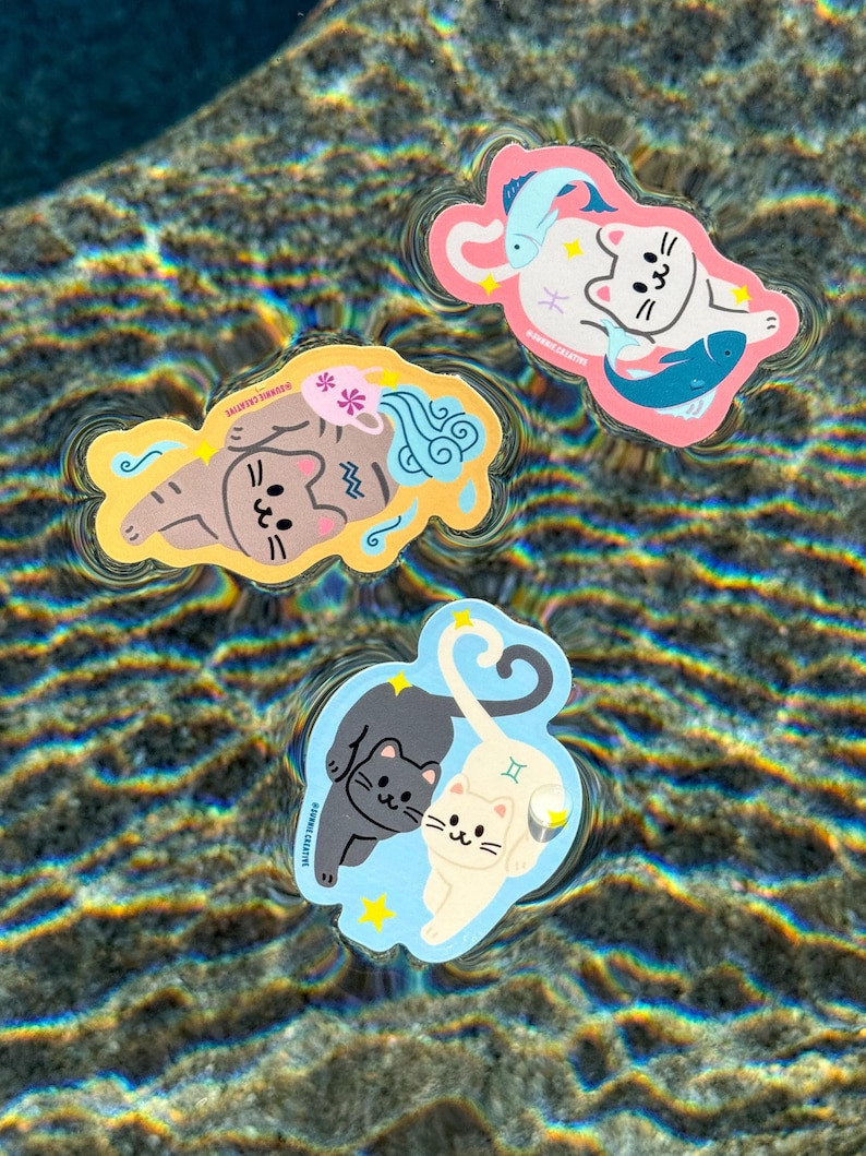 May include: Three colorful cat-themed stickers floating in water. One sticker features a brown cat with blue water-like designs and pink flowers. Another shows a white cat with two blue fish. The third sticker has two cats, one black and one white, with a blue background.