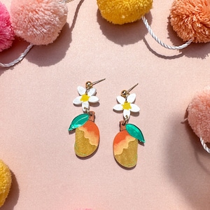 May include: A pair of gold earrings with a mango, flower, and green leaf design. The earrings are on a pink background with colorful pom-poms.