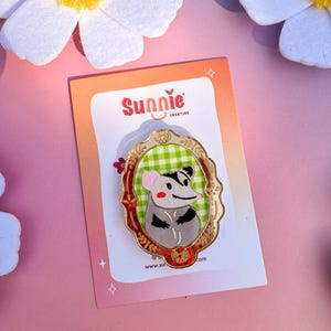 May include: A gold-colored enamel pin featuring a cartoon opossum wearing a green and white checkered shirt, framed in a gold oval with red and green accents. The pin says "Sunnie Creative" and "www.sunnie.com".