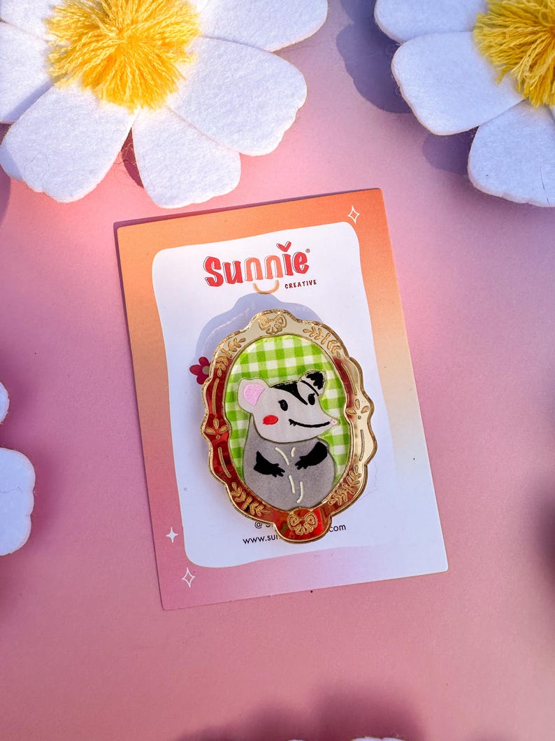 May include: A gold-colored enamel pin featuring a cartoon opossum wearing a green and white checkered shirt, framed in a gold oval with red and green accents. The pin says "Sunnie Creative" and "www.sunnie.com".