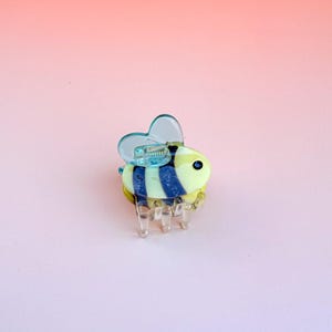 May include: A bee-shaped hair clip made of clear plastic. The bee has a yellow body with blue stripes, blue wings, and clear legs. The clip is set against a gradient pink and peach background.