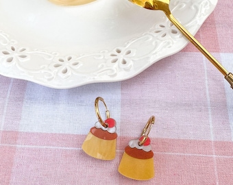 Peut inclure: Boucles d'oreilles créoles dorées avec un motif de dessert. Chaque boucle d'oreille représente un petit dessert à la crème anglaise jaune avec un dessus marron, de la crème blanche et une cerise rouge. Fond rose et blanc à carreaux.