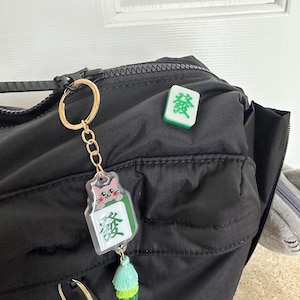 May include: A black backpack with a gold keychain featuring a green mahjong tile with a cartoon cat and a green tassel. A separate green mahjong tile with the Chinese character "發" is attached to the backpack.