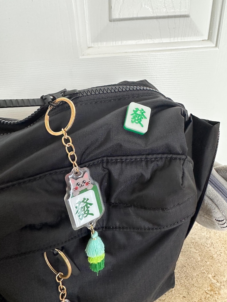 May include: A black backpack with a gold keychain featuring a green mahjong tile with a cartoon cat and a green tassel. A separate green mahjong tile with the Chinese character "發" is attached to the backpack.
