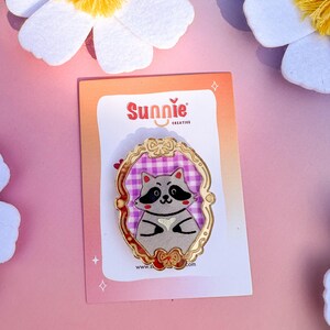 May include: A gold-colored enamel pin featuring a cartoon raccoon wearing a pink and white checkered dress. The raccoon is holding a heart in its paws. The pin is surrounded by a gold-colored frame with a bow at the bottom. The pin is on a white card with the text "Sunnie Creative" and a website address.
