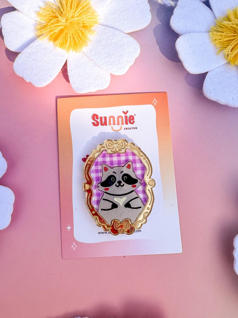 May include: A gold-colored enamel pin featuring a cartoon raccoon wearing a pink and white checkered dress. The raccoon is holding a heart in its paws. The pin is surrounded by a gold-colored frame with a bow at the bottom. The pin is on a white card with the text "Sunnie Creative" and a website address.