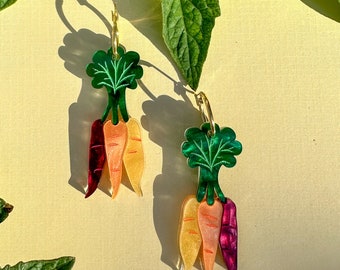 Peut inclure: Une paire de boucles d'oreilles créoles dorées avec un groupe de trois breloques en forme de carottes. Les carottes sont orange, jaune et rouge avec des sommets verts.