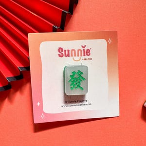 May include: A green and silver mahjong tile with the Chinese character for "prosperity" on it. The tile is on a white card with the text "Sunnie Creative" and a website address.