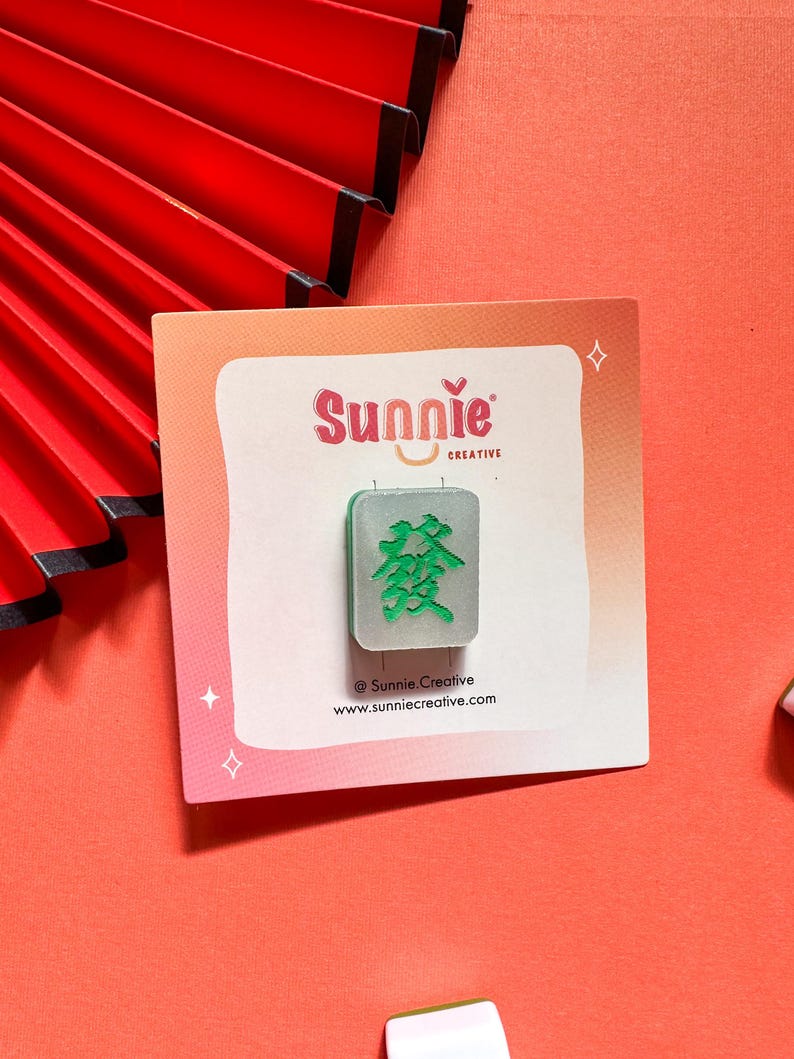 May include: A green and silver mahjong tile with the Chinese character for "prosperity" on it. The tile is on a white card with the text "Sunnie Creative" and a website address.