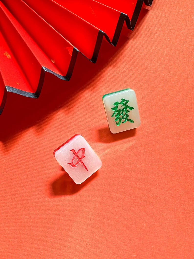 May include: Two white mahjong tiles with Chinese characters on a red background. The tile on the left has a red character, and the tile on the right has a green character.
