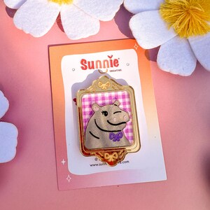 May include: A gold-colored enamel pin featuring a cartoon hippopotamus wearing a purple bow tie. The hippo is framed in a gold-colored frame with a pink and white checkered background. The pin is on a pink and white card with the text "Sunnie Creative" and "www.sunnie.com".