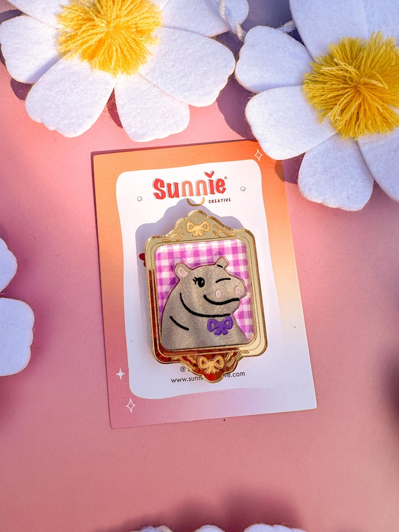 May include: A gold-colored enamel pin featuring a cartoon hippopotamus wearing a purple bow tie. The hippo is framed in a gold-colored frame with a pink and white checkered background. The pin is on a pink and white card with the text "Sunnie Creative" and "www.sunnie.com".