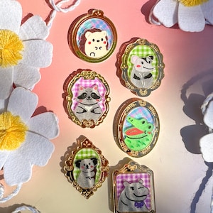 May include: Set of seven gold-framed pins featuring various animals, including a hedgehog, raccoon, crocodile, koala, and a hippopotamus. Each pin has a checkered background and is surrounded by a decorative gold frame.