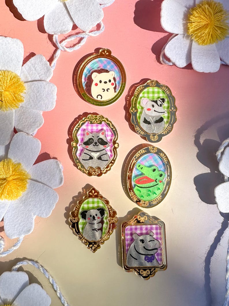 May include: Set of seven gold-framed pins featuring various animals, including a hedgehog, raccoon, crocodile, koala, and a hippopotamus. Each pin has a checkered background and is surrounded by a decorative gold frame.
