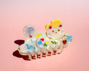 May include: A white and iridescent cat-shaped hair claw clip with a yellow hat and colorful accents. The claw clip features a playful design with a question mark, swirls, and a palette of colors. The clip is designed to hold hair in place.