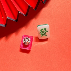 May include: Two small square pins with a red and white background. One pin features a green Chinese character, the other pin has a silver pin back.